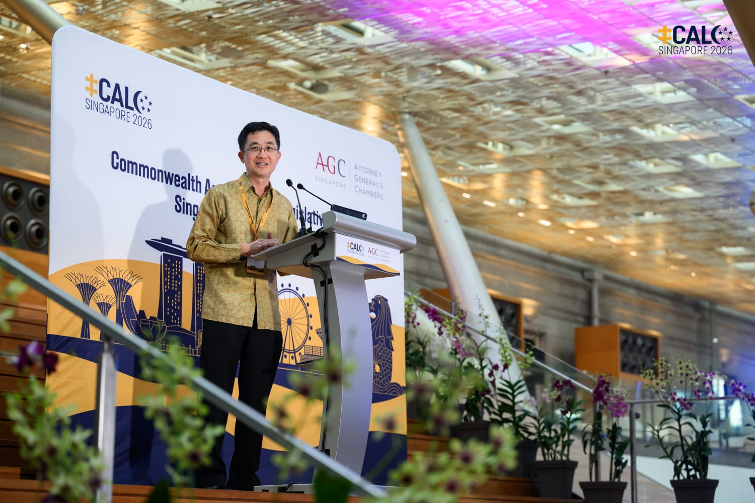 Mr Lionel Yee SC, Deputy Attorney-General of Singapore, in a gold batik shirt delivers a speech at a podium at the Commonwealth Association of Legislative Counsel Singapore Conference 2026, co-organised with AGC Singapore, in a venue decorated with orchids.